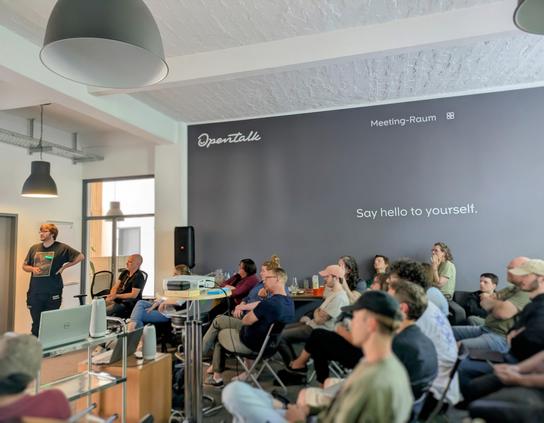Ein Team sitzt aufmerksam in einem modernen Meetingraum von OpenTalk, während ein Kollege vorne eine Präsentation hält. An der Wand steht der Schriftzug „Say hello to yourself.“.