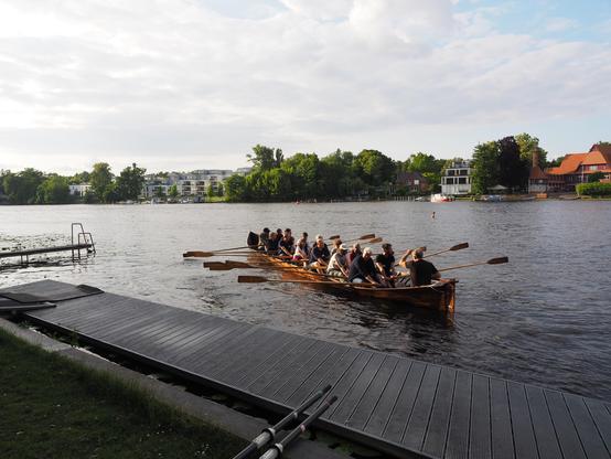 Eine Gruppe von Menschen rudert gemeinsam in einem traditionellen Holzboot auf einem ruhigen Fluss, umgeben von grünen Bäumen und Uferhäusern.