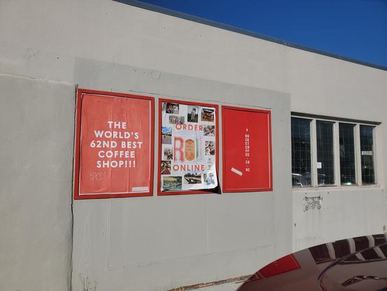 Pale wall, blue sky. Three posters on wall, all with red frames furthest is red and middle is mostly white, both with unintelligible writing. Nearest is red background, white text stating 'The World's 62nd best coffee shop!!!'
