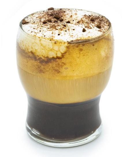 A glass of layered coffee drink with foam and chocolate sprinkles on top.