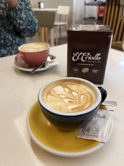Two cups of coffee with latte art are on a white table, next to a box of "El Criollo" coffee. The first cup is in a pink mug, while the second one is in a blue mug with a yellow saucer. A sugar packet is next to the second cup.