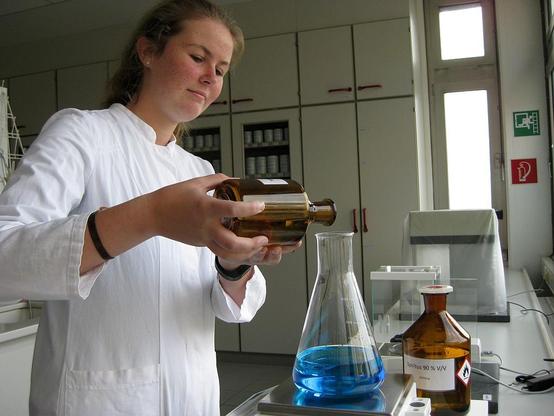 Im Labor: Eine PTA-Schülerin in weißem Kittel schüttet aus einer braunen Flasche eine Flüssigkeit in einen Kolben, der schon grün-blaue Flüssigkeit enthält. Der Messkolben steht auf einer Waage. Daneben auch noch eine Braune Flasche mit 90-prozentigem Spiritus.