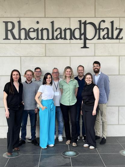 Gruppenfoto von neun Teilnehmenden der KNOER-Tagung 2025 vor der Wand mit der Aufschrift „Rheinland-Pfalz“ an der Vertretung des Landes beim Bund in Berlin.