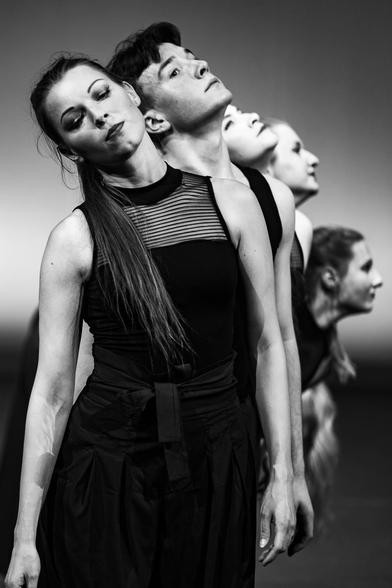 The image showcases a line of dancers, each adorned in dark attire. They are captured in a monochrome setting of a performance space. In the foreground, the focus is on the lead dancer, with the others receding into the background.