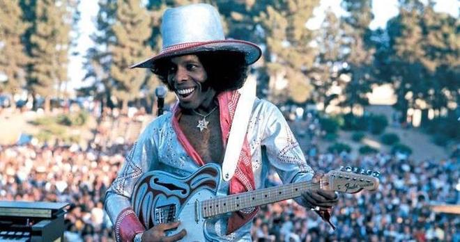 A photo of Sly Stone playing guitar at a live outdoor performance.