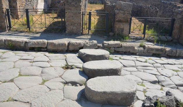 Foto von Ausgrabungen in Pompeji. Erhöhte Steinquader sind in der Mitte einer antiken Straße zu sehen. Sie ermöglichten auch bei regennassen Straßen den bequemen Übergang von einer Straßenseite zur anderen. Der Abstand zwischen den Steinen wurde so bemessen, dass die Fuhrwerke ungehindert mit ihren Rädern passieren konnten.