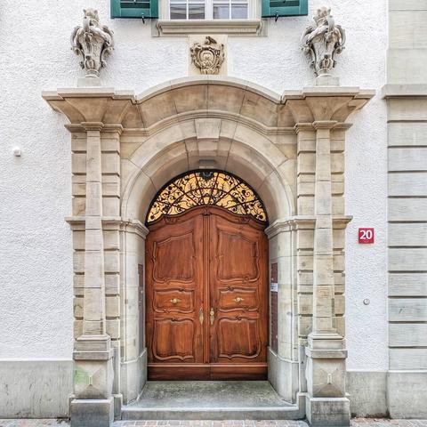 A grand, ornate Baroque-style wooden double door with a rich, warm brown finish. The door is set within a large, arched stone portal, flanked by two thick, fluted pilasters topped with decorative corbels featuring sculpted elements. Above the arch, a central coat of arms or crest is carved into the stone. The semicircular transom above the door is filled with intricate black wrought-iron scrollwork set against a golden background, allowing light to filter through. To the right of the door, a red rectangular plaque with the number "20" is affixed to the plain white wall. Above the portal, a window with green louvered shutters is visible. The ground in front of the door is made of light-colored pavers.