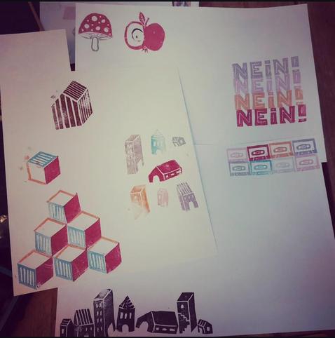 Mehrere Blätter Papier, auf denen verschiedene Stempel getestet wurden: unterschiedliche Häuser, ein Rautenmuster, ein Pilz, ein Apfel, das Wort "Nein" und Kassetten.