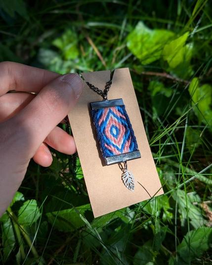 Photo d'un pendentif porte-clés tissé aux couleurs du drapeau trans sur une cartonnette de présentation tenue par une main sur fond d'herbe et de lierre.