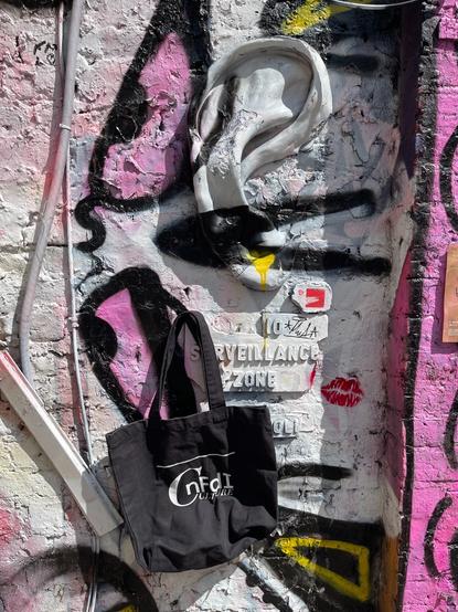 Die NFDI4Culture-Tasche hängt an einer grau, schwarz, rosa und gelb gestalteten Wand mit einem plastisch gestalteten Ohr unter dem auf einem Schild steht: Surveillance Zone.
London, Shoreditch
CC0
Foto: Martin Albrecht-Hohmaier