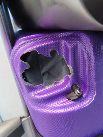 Detail of one of the wheels ripped off of a new hardshell suitcase, which ripped off part of the shell itself, exposing the lining of the suitcase. There's a metal screw pointing out where the wheel once connected.