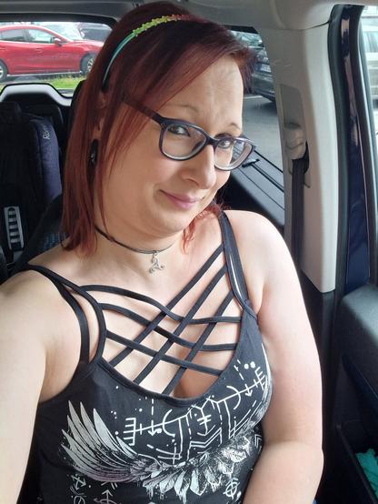 Das Bild zeigt eine Frau mit roten Haaren, die in einem Auto sitzt. Sie trägt eine schwarze Top mit einem auffälligen Strappy-Design und einem weißen Druck, der stilisierte Flügel und geometrische Muster zeigt. Um ihren Hals hat sie eine schwarze Kette mit einem kleinen Anhänger. Sie trägt eine Brille mit dunkelblauen Rahmen und ein buntes Haarband. Der Hintergrund zeigt die Innenausstattung des Autos, einschließlich der Beifahrerseite und des Fensters, durch das ein rotes Auto im Parkhaus zu sehen ist.