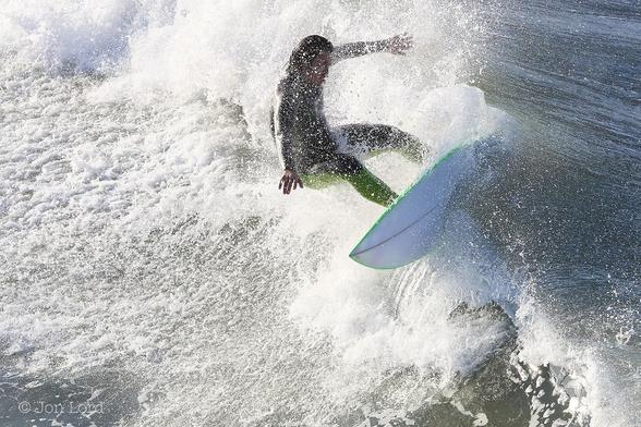 A Colour Water Sports Photo In Landscape Format. Above The Centre Is A Male Surfer Surrounded By Spray And Surfing Along The Crest Of A Breaking Wave. The Sea Is To The Right While The Shore To The Left. The Surfer Is Coming Towards The Camera And Moving Slightly To The Left. The Board Is At A Steep Angle Of Sixty Degrees Of More With The Surfers Legs Firmly On The Board But With His Body Nearly Vertical And His Arms Outstretched As He Maintains His Balance. His Board Is White With A Thin Green Stripe Along The Edges. The Forward Half Of The Board Is Clear Of The Water While The Aft Is Imbedded In The Wave. The Surfer Has Long Dark Hair And Is Wearing A Black Wetsuit With Green Coloured Legs. 
The Location: Hermosa Beach, California (2014).