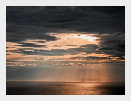 The photo is of a dramatic seascape near sunset. At the top of the photo, dark, heavy clouds take up the upper portion of the frame. Underneath them there is a lower layer of lighter, more wispy clouds.  Neat the centre, there is a hole in the clouds. Through it bright sunlight is partially visible. From the break there are golden rays casted down toward a calm, dark ocean. The ocean is at the very bottom of the frame and it mostly dark and moody except for the right hand side which is illuminated by the sun rays.