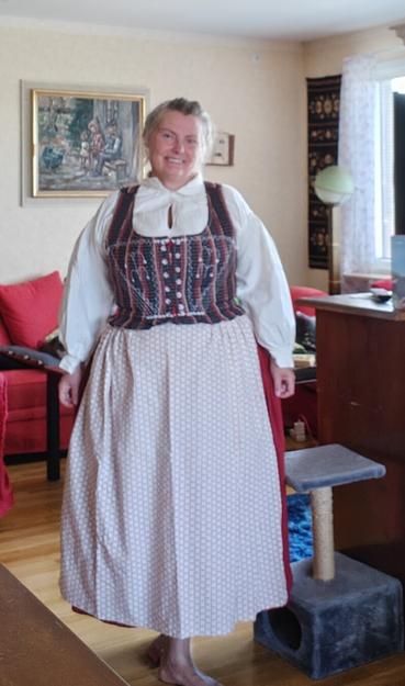 A white middle aged woman is standing in a living room. Behind her are a red armchair and sofa. Beside her is a small pillar with a platform for a cat, covered in grey shaggy fabric, and rope, and a brown cupboard. She is wearing a a white blouse with wide sleeves gathered at the cuffs, a red skirt, which is hardly visible behind an apron, and a tight bodice from a dark checked fabric. The apron is cream white with a small overall print in red. She is barefoot.