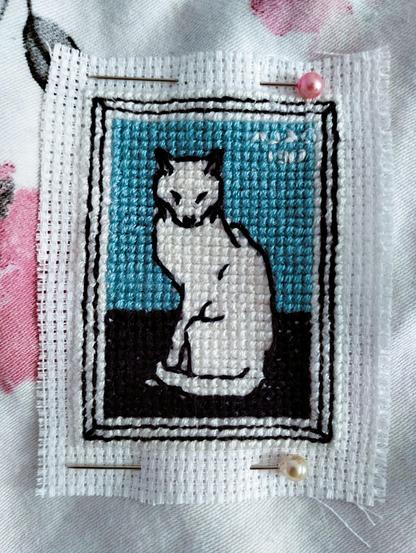 Julie de Graag "Sitting Cat".  Wersja haftowana krzyżykami, miniaturowa (28x37 krzyżyków), siłą rzeczy, uproszczona.
Biały kot siedzi na granatowym stole (?), a za nim jest niebieskie tło. Wyraźne kontury, bo jeśli dobrze pamiętam, oryginał był litografią.