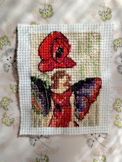 CIcely Mary Barker "Poppy Fairy".  Wersja haftowana krzyżykami, miniaturowa (28x37 krzyżyków), siłą rzeczy, uproszczona.
Wróżka ubrana na czerwono, z czerwono-fioletowymi skrzydełkami i brązowymi włosami, stoi obok maku. Jest od kwiatka nieco niższa.