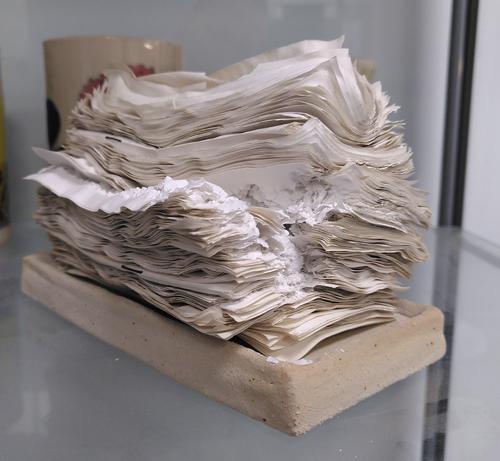 A pile of falling apart stack of old magazines pages after they went into a ceramic kiln at a thousand degrees celsius. It looks very light, we can still see all the individual pages, the ink has burnt out so the pages are all off white beige.