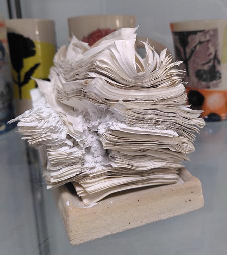 A pile of falling apart stack of old magazines pages after they went into a ceramic kiln at a thousand degrees celsius. It looks very light, we can still see all the individual pages, the ink has burnt out so the pages are all off white beige.