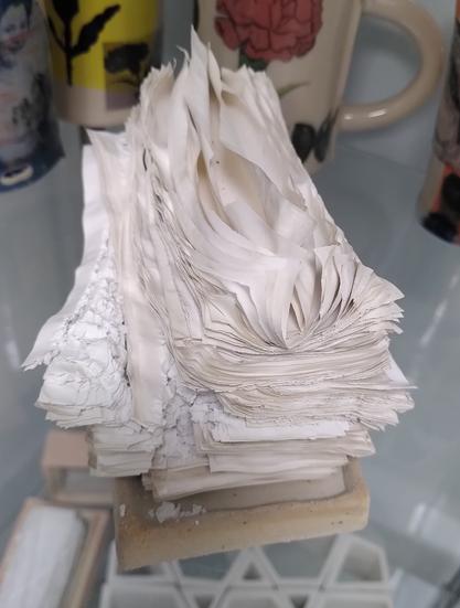 A pile of falling apart stack of old magazines pages after they went into a ceramic kiln at a thousand degrees celsius. It looks very light, we can still see all the individual pages, the ink has burnt out so the pages are all off white beige.
