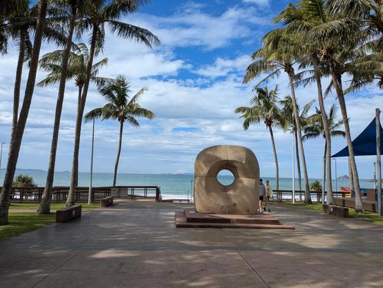 A wide walkway lined with Palm trees leading to the beach. In the middle of the walkway is a sculpture with a hole calved in the middle.