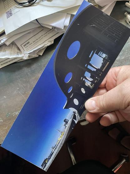 A hand holds a postcard in panoramic format. Most of the picture is filled with an azure blue, cloudless sky. On the right, the picture is dominated by a curved concrete structure on pillars, the structure is pierced by circular holes. It is a solar sail on the roof of a building. The roofs of urban buildings can be seen at the bottom of the picture