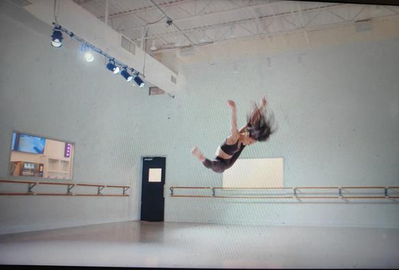 Une danseuse fait un grand écart facial en sautant et en baissant la tête dans un studio de danse (photo moche de mon écran). Ses cheveux volent mais enfin surtout c'est inhumain.