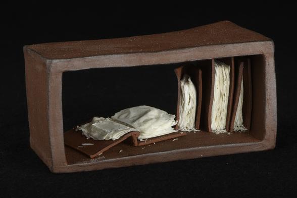 A mini rectangle book shelf opened front and back. It has 3 mini books standing vertical and one flat opened. In each mini book there are burn pages of paper. The book shelf is made of terracotta clay and the book covers are terracotta paperclay fired at 1220 degrees celsius. The paper pages have been added afterwards and then the piece has been fired again at a 1000 degrees celsius.