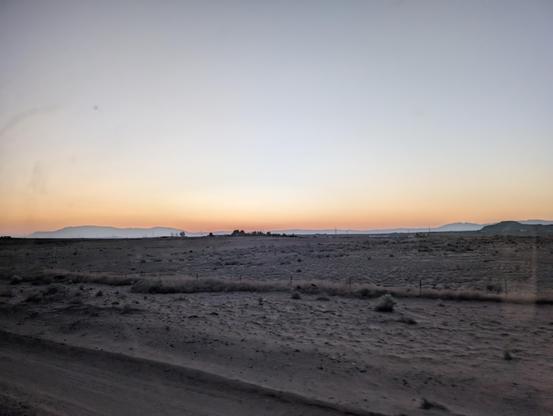 Photo of New Mexico sunrise over a desert. Sandy dunes and distant hills, slight orange tint across the horizon. The sky seems very pale shade of blue