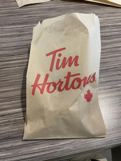 A paper bag on a table with the "Tim Hortons" logo in red and a maple leaf. The setting conveys a casual, everyday coffee shop vibe.