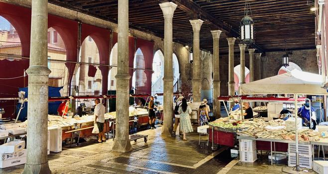 The photo shows the fish market of Venice at the Mercato di Rialto, Campo de la Pescaria. Stalls filled with fresh fish and seafood are lined up beneath a high wooden-beamed roof supported by thick stone columns. Red fabric shades hang between the arches, casting a warm glow. The wet stone floor reflects the light. Shoppers in summer clothes and hats wander between the vendors. Old lanterns hang from the ceiling, adding to the historic, vibrant atmosphere.