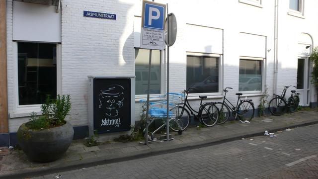 straat met witte muur van wonigen/pand met plantenbak en parkeerbord, geparkeerde fietsen, winkelwagen en een bord zwart gemaakt met portret erop en : 'Skinny!'