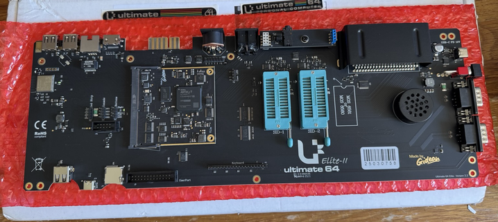 A top-down photo of a motherboard populated with various components including a pair of ZIF sockets, connector headers, various ports along the edges. Main PCB colour is black and is sparsely populated. Legend printed on the board reads Ultimate 64 Elite-2 with “made by Gideon” also featuring