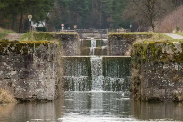 The image depicts a waterway with multiple small waterfalls or weirs. The structure appears to be made of stone or concrete and is surrounded by greenery, including trees in the background. There is a calm water surface reflecting the scenery, and you can see a stone bridge in the distance. The overall setting suggests a tranquil natural environment, possibly in a park or rural area.

Image Credits: Wikimedia / Derzno / CC BY-SA 4.0