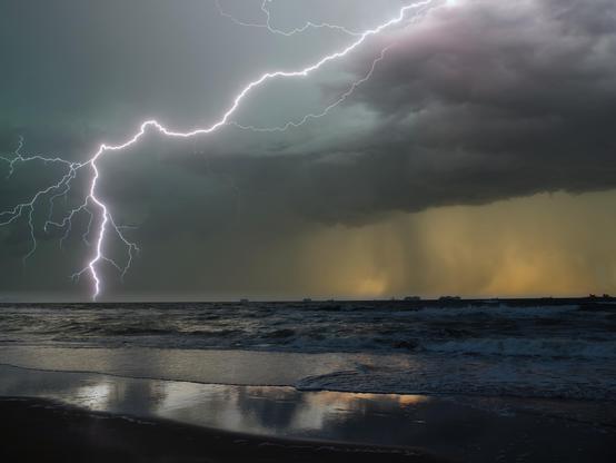 A sandy beach in the evening, with dark waves breaking. The sea is rough, the sky darkened by threatening clouds, from which a powerful flash of lightning strikes across the sky. Ships are visible on the horizon, some of which have lights. In the distance, the first rain clouds are forming long veils.