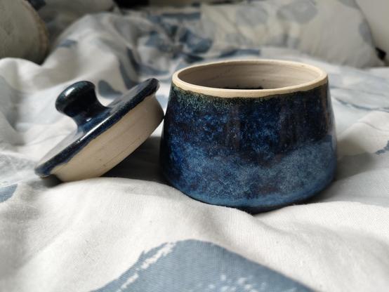 a round small jar with the lid next to the body of the jar, on top of a white duvet with blue patterns. The body of the jar is slightly wider at the base than the rim. The jar is glazed in a deep blue with a lot of variation in the glaze. It is lighter at the bottom and darker on top. The lid has a round knob and is is glazed the same. Both have bare clay where the lid and body overlap, without any glaze - around the rim and top of the inside of the body, and around the bottom of the lid.