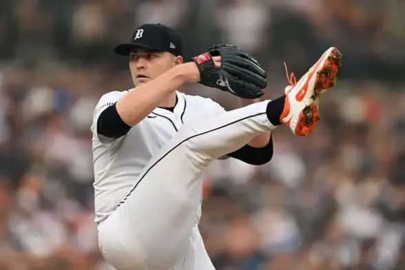 A baseball pitcher in a white uniform prepares to throw, lifting one leg high with a glove on one hand and a focused posture.