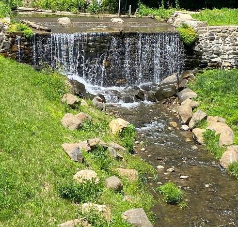 In the distance (upper picture), water from a relatively small creek flows over a stone wall some 4 feet tall, splashing on larger rocks below (center pic) ... after the fall, the creek is much thinner and flows down to the bottom right corner of the picture.  At this point, lighter colored stones sit on green grass on either side of the creek.