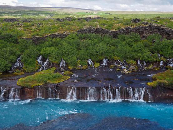 A long horizontal series of two-tiered waterfalls coming out of a lava field that's covered by moss and low bushes and falling into a blue-green river.