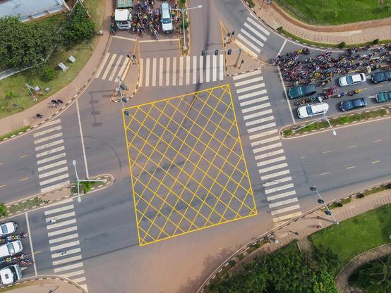 Aerial view of a busy intersection in Kigali, Rwanda, showing a large crossroads with white striped crosswalks on all four sides. The center of the intersection is overlaid with a yellow grid pattern. Heavy traffic is visible with numerous cars lined up on the right side of the image, while crowds of people can be seen gathered near vehicles in the upper portion. The intersection is surrounded by paved areas, green spaces, and landscaping, illustrating the urban infrastructure improvements mentioned in Rwanda's junction upgrade plans.