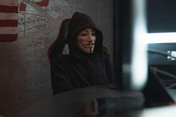 Dans une pièce, une personne est assise sur un fauteuil. Elle porte un hoodie, avec la capuche sur la tête. Son visage est caché par un masque de type Casa de Papel ou Anonymous.