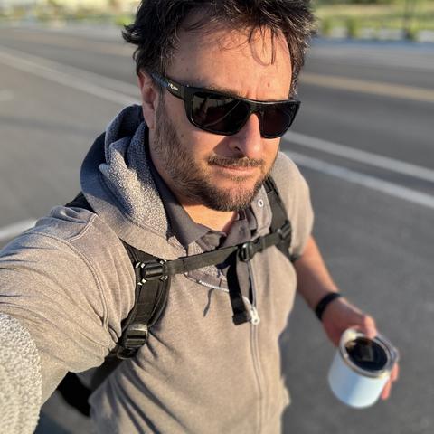 Selfie of a man walking down the street holding a cup of coffee. He has short, spiky hair and is wearing sunglasses, a hoodie, and backpack.