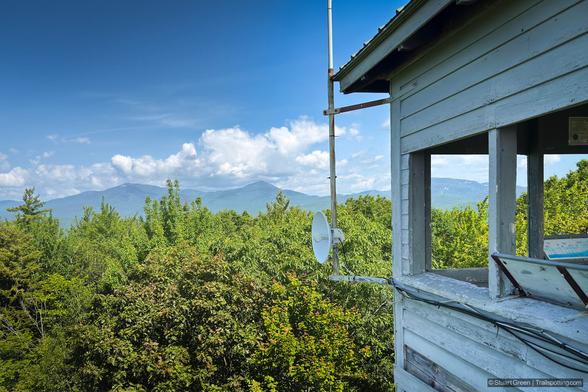 A scenic view from a wooden lookout tower positioned on the right side of the frame. The structure is built of aged, weathered timber with visible grain and knots, evoking a rustic, utilitarian design. A rectangular window opening in the tower offers a clear view of a densely forested valley filled with vibrant green treetops. Beyond the forest, layers of misty blue-gray mountains stretch into the distance, softening as they recede into the horizon.
