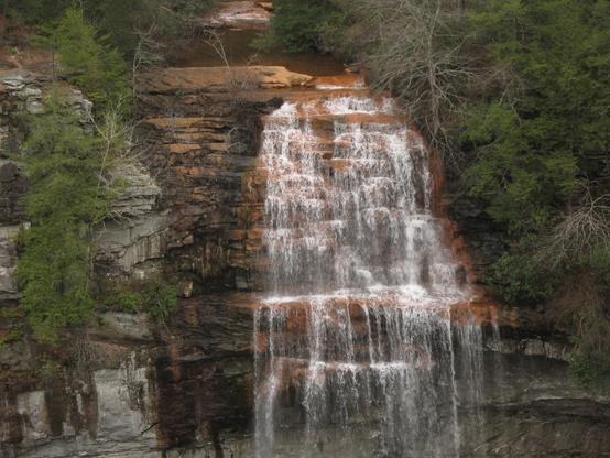A waterfall cascades over a rugged, reddish-brown rock face surrounded by evergreen trees and bare deciduous branches. The water flows in multiple streams, spreading out across the uneven surface of the cliff before plunging down the rock wall. The reddish hue of the rock suggests iron-rich minerals, giving the waterfall a distinctive appearance. The setting appears natural and secluded, with a mix of forest and exposed stone.
