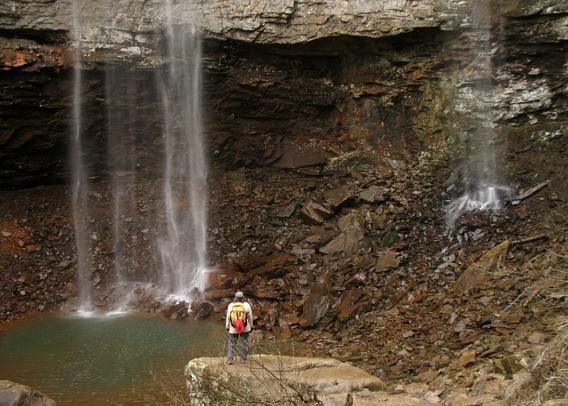 A hiker wearing a yellow backpack stands on a rock ledge, facing a rocky cliff where several thin streams of water cascade down into a small, greenish pool below. The waterfall is delicate and appears to trickle over the edge of a high rock shelf. The surrounding area is rugged, with loose stones and stratified rock layers.