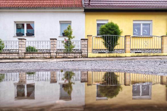Das Bild zeigt die Vorderseiten von zwei Reihenhäusern mit zwei verschiedenen Fassadenfarben: Weiß und Gelb. Die Häuser haben leicht unterschiedlich rote Ziegeldächer und jeweils ein Doppelfenster und ein einzelnes Fenster, die wie spiegelverkehrt angeordnet sind. Vor jedem Haus befindet sich ein Zaun aus Stein und Metall mit dekorativen Elementen. Entlang des Zauns wachsen mehrere Pflanzen und Blumen, die etwas Farbe und Lebendigkeit hinzufügen. Im Vordergrund des Bildes liegt eine Pfütze auf der Straße, die eine klare Spiegelung des Hauses und des Zauns zeigt.