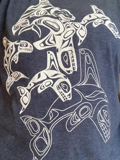 A dark blue tshirt with white design of many orcas, in a northwest (North America) Indigenous art style. Most of them are drawn with very thick white lines, but the largest one at the bottom is drawn with thin lines. Most of them are facing right, but one in the middle is facing left and down as if diving deep.