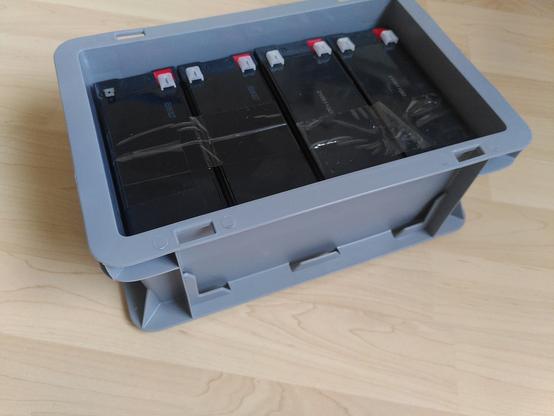 Grey standard 30x20 cm euro box with 4 black lead acid batteries inside. The batteries fit snugly into the box with about a millimeter of tollerance.
The box is placed on a brightly lit wooden floor.