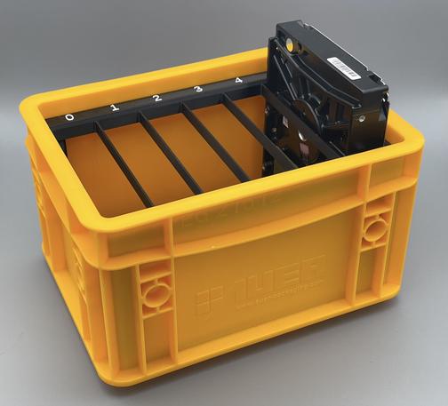 A yellow Eurobox in cute 15×20 cm size. Inside the box is a 3d-printed black frame with 6 equally sized openings to fit hard drives. There are white labels with numbers 0 to 5 on each of the compartments. The right most, with number 5 is filled with a 3.5” hard drive.
