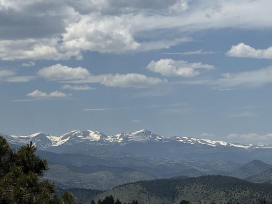 Snow capped Mountains rise in the distance over hill in various shades of green. The sky has scattered white clouds and is a light blue